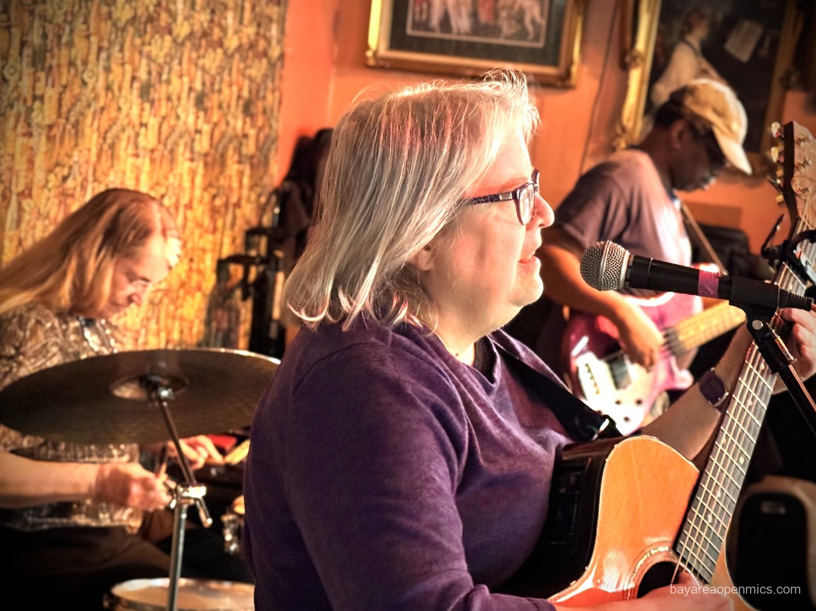 A woman sings in the foreground, another plays drumkit, and a man plays bass in the background