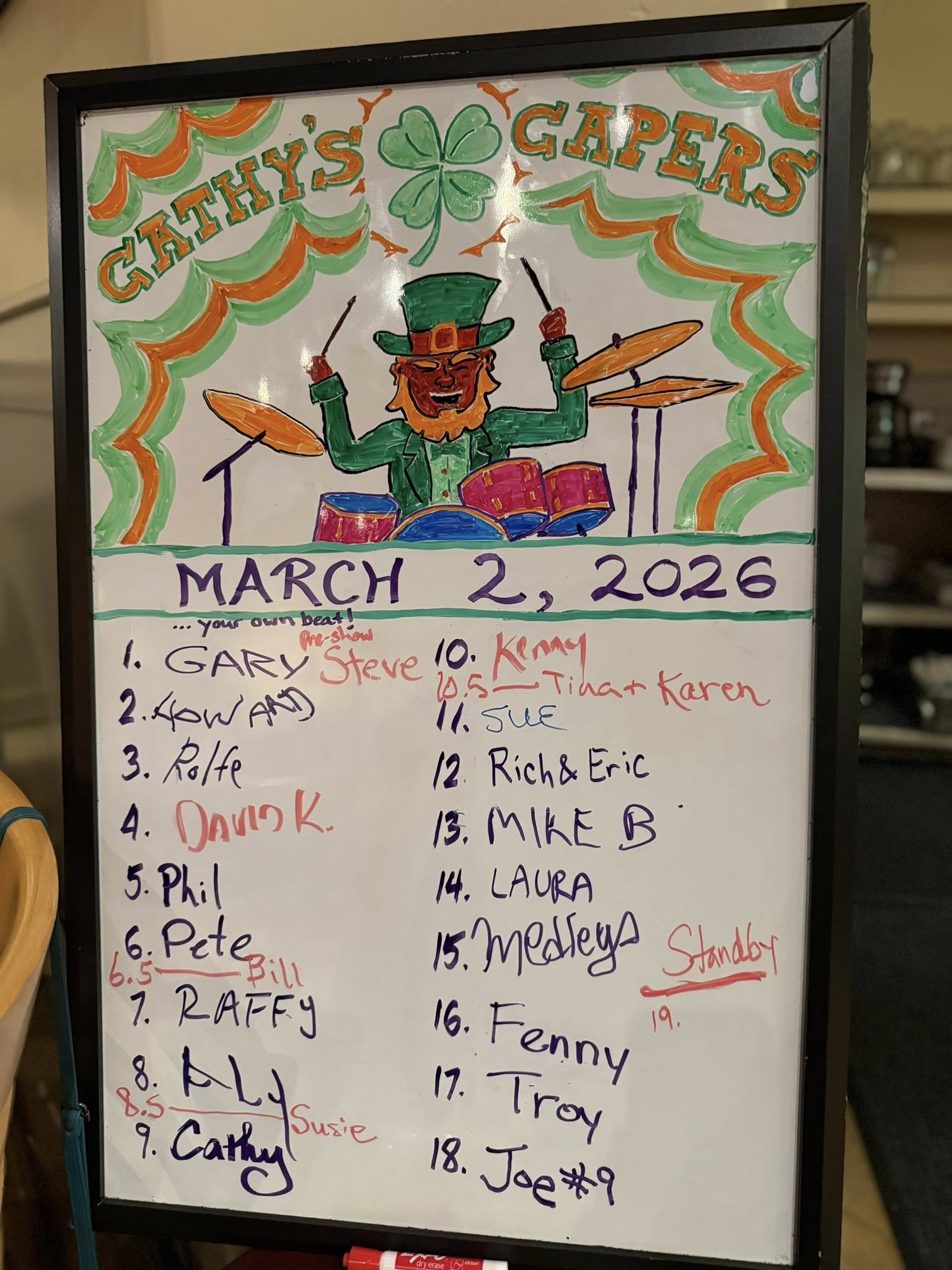 a colorful white board depicts a leprechaun playing a drum kit and declares "Cathy's Capers March 2, 2026" above a list of open mic performer's names