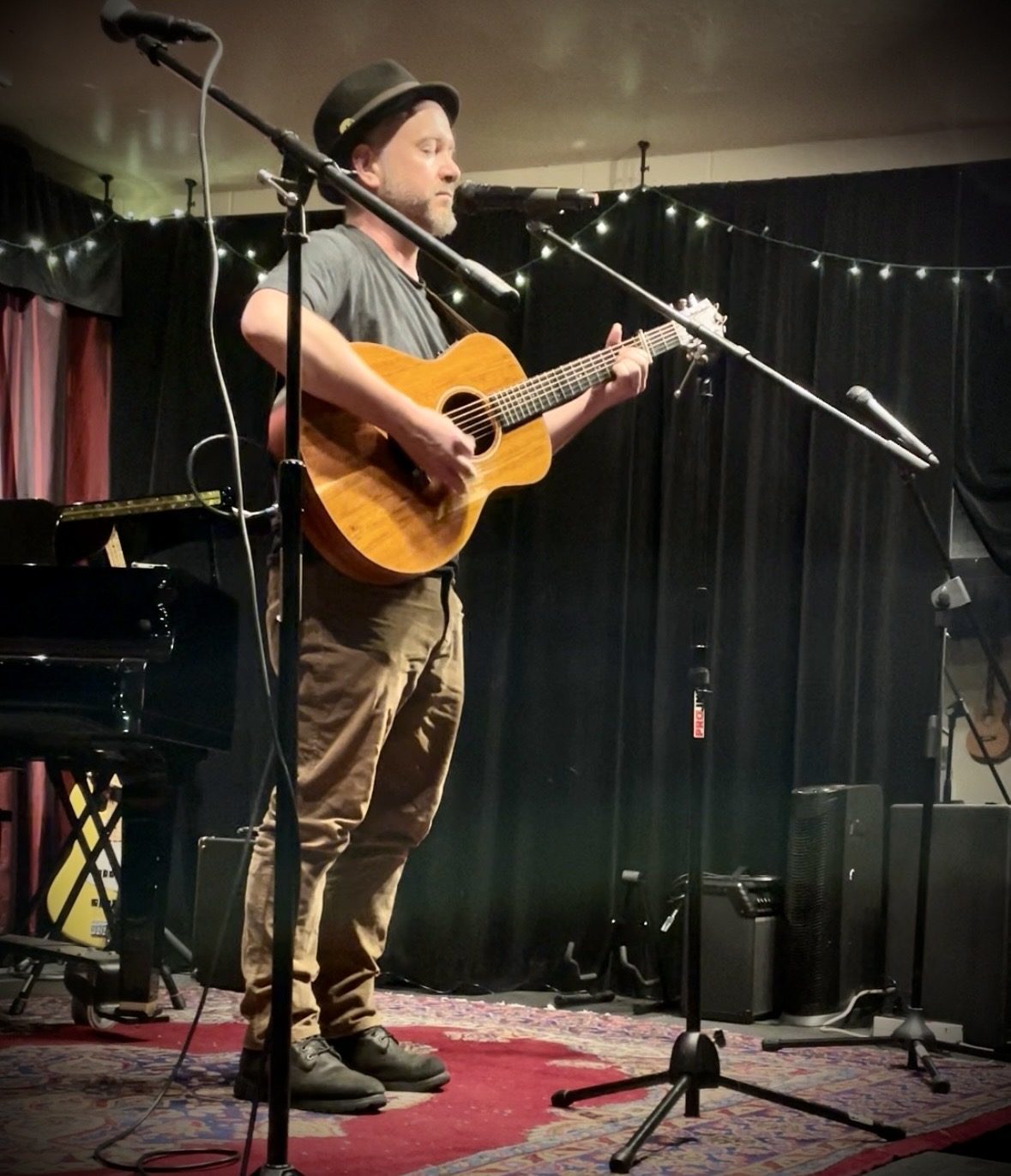 A man in a stingy-brim dark fedora, t-shirt, and courderoy pants plays acoustic guitar while singing into a microphone onstage