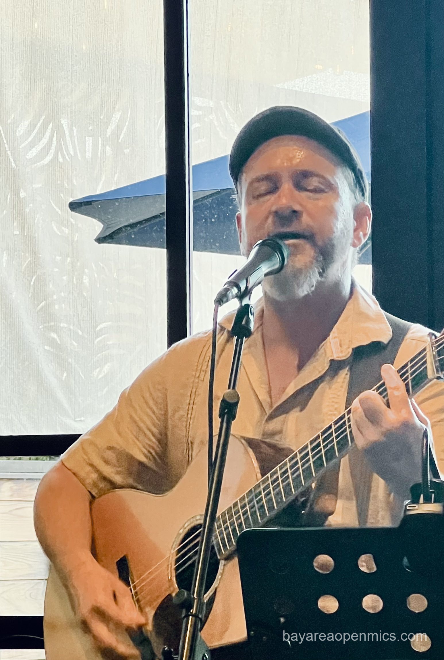 a man in an Irish flat cap plays guitar and sings in front of a mic
