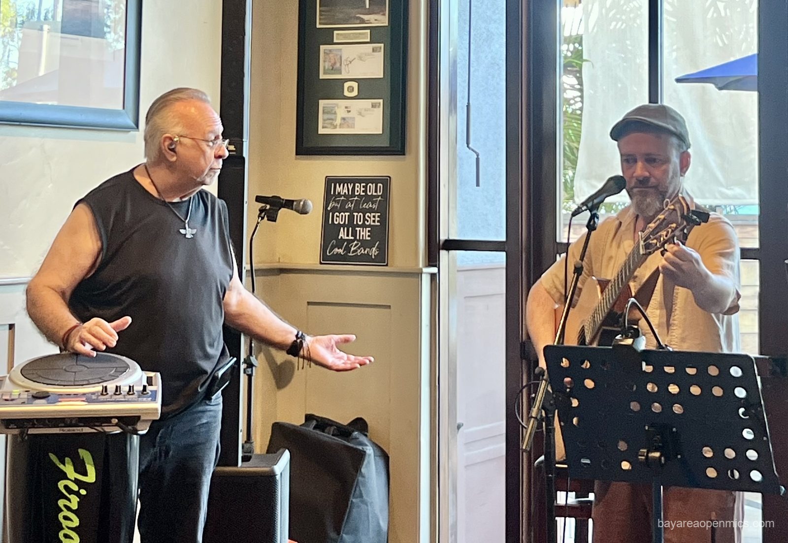 a man in a black sleeveless tee and a zoroastrian amulet gestures plaintively toward a man opposite him tuning an acoustic guitar