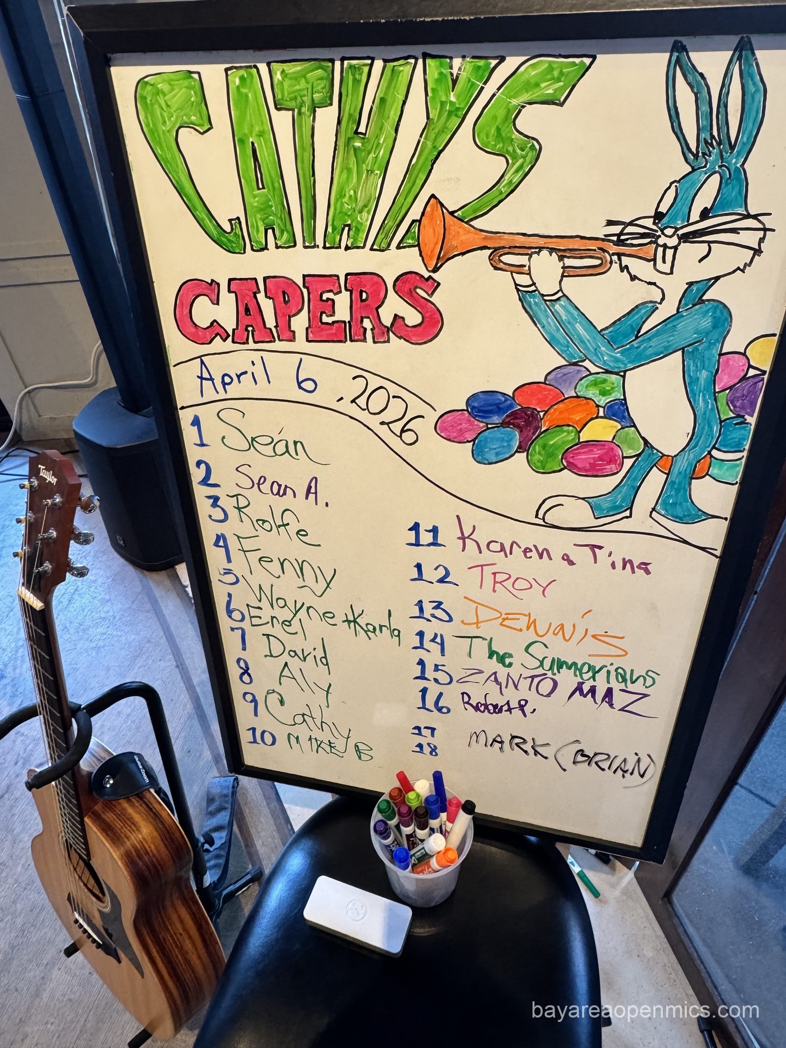 a colorful white board depicts Bugs Bunny playing a trumpet next to a pile of Easter eggs and declares "Cathy Capers April 6, 2026" above a list of open mic performer's names