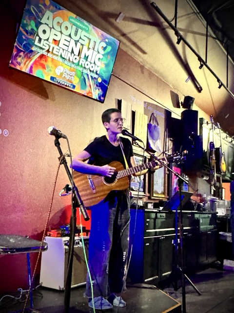 A woman with a buzz cut and baggy jeans performs onstage beneath the Acoustic Open Mic Listening Room sign at Pinot's Palette