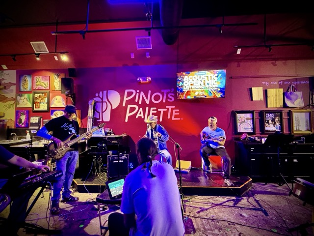Accompanists are depicted at Pinot's Palette Open mic: an arm extends into the frame over a piano keyboard; a bass player in a baseball cap, jeans and sandals plays while standing; a lead singer in a cowboy hat holds a harmonica; an electric lead guitar player with short white hair and a pair of sunglasses on his head jams along to the singer's left onstage, and in the foreground we can see open mic host John Iacono's with his back turned to the camera, a glowing iPad with his sound controls in front of him, and he is slightly bent forward, sitting atop te cajon he is playing