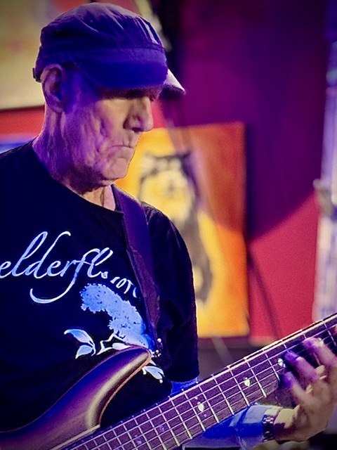 An older man in a snug cloth baseball cap plays a six-string bass guitar while wearing a shirt that reads, 'Elderflower.' at Pinot's Palette open mic in Livermore