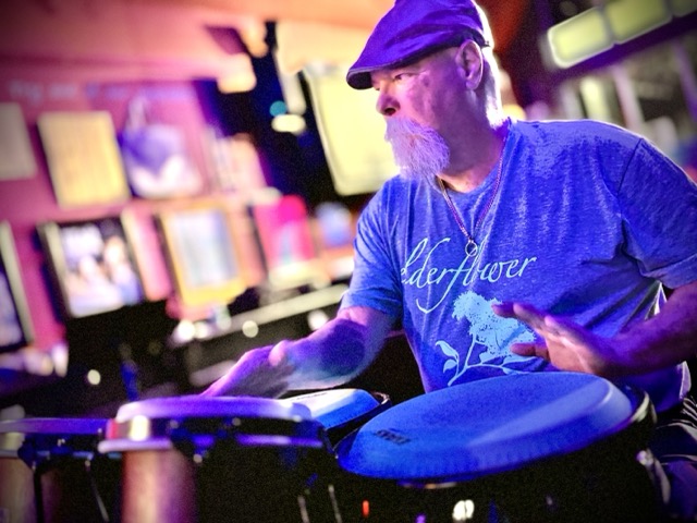 A bearded man in a flat cap expertly plays congas with bongos in the foreground at Pinot's Palette