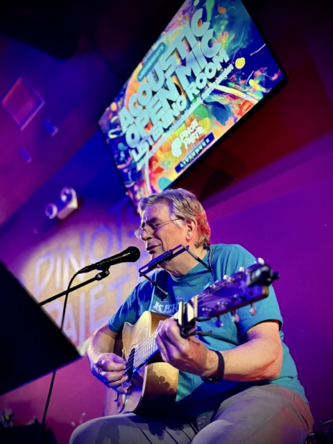 Richard Lappin with a harmonica bracket around his neck sings while playing acoustic guitar beneath the Acoustic Open Mic Listening Room sign