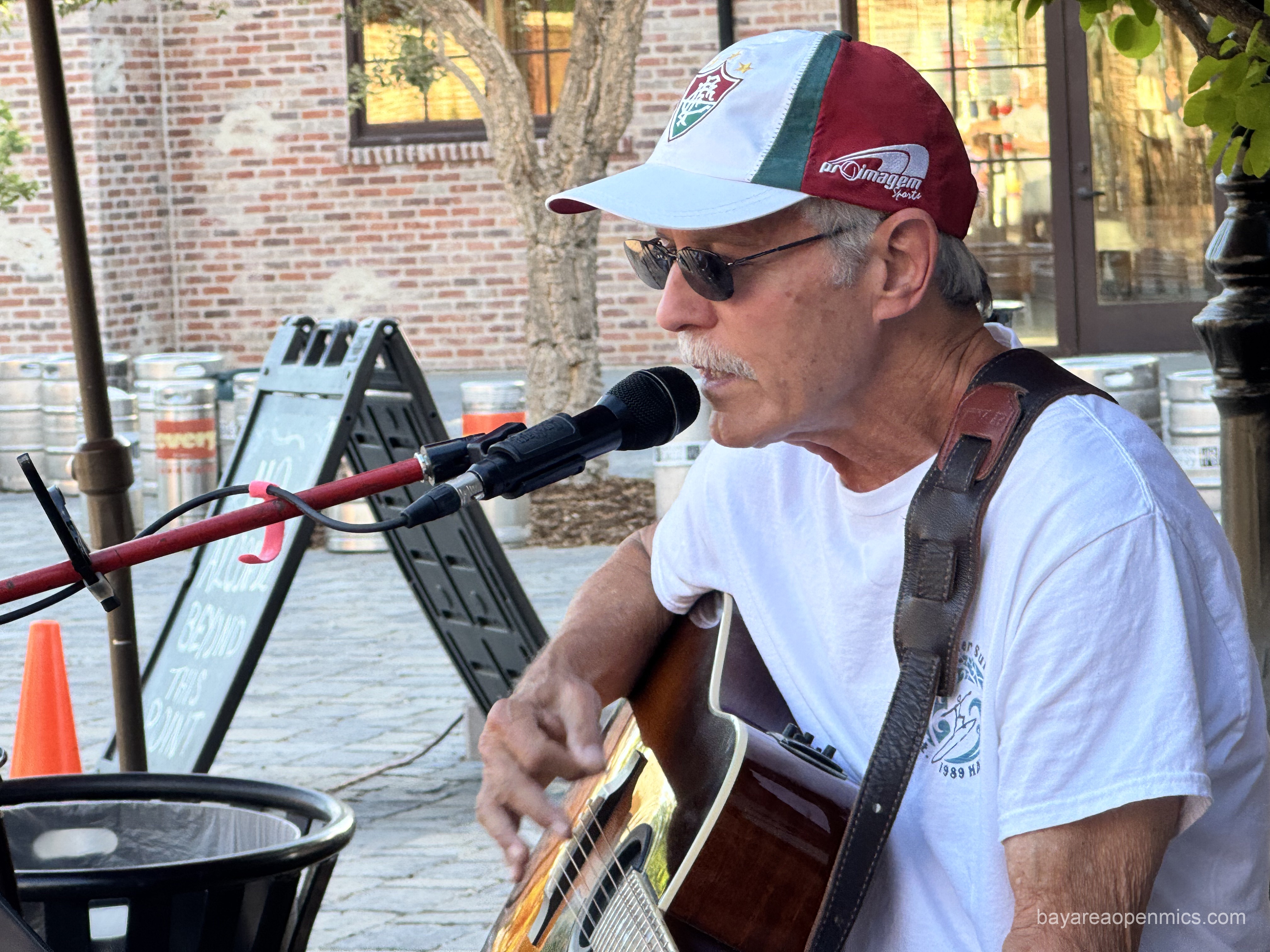 a man with a grey mustache in a baseball cap and sunglasses strums a guitar while singing into a microphone 