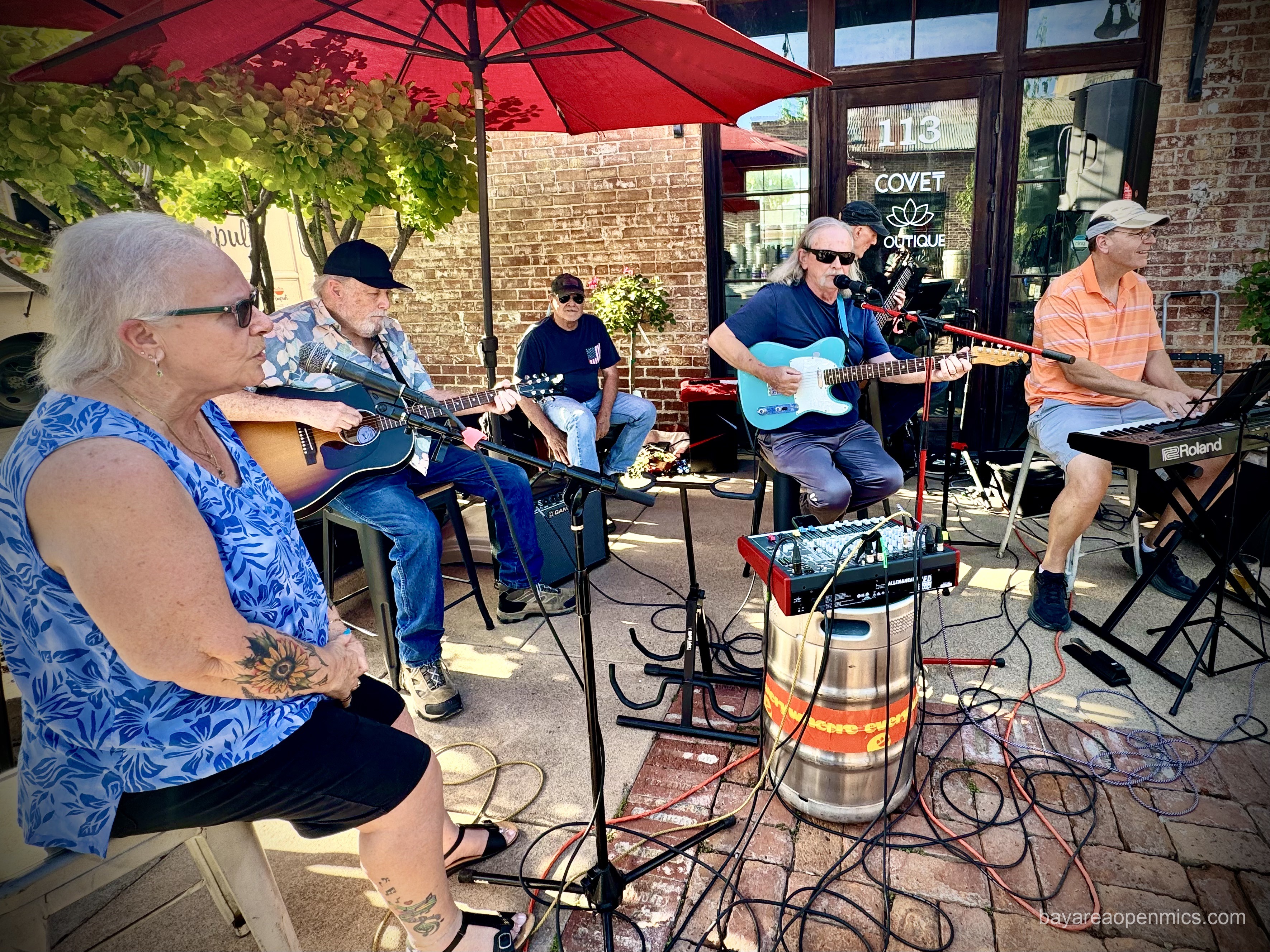 A mixing board is perched on a steel beer keg in the midst of six older rock musicians gathered around an assembly of microphones and cords under a large red sun umbrella