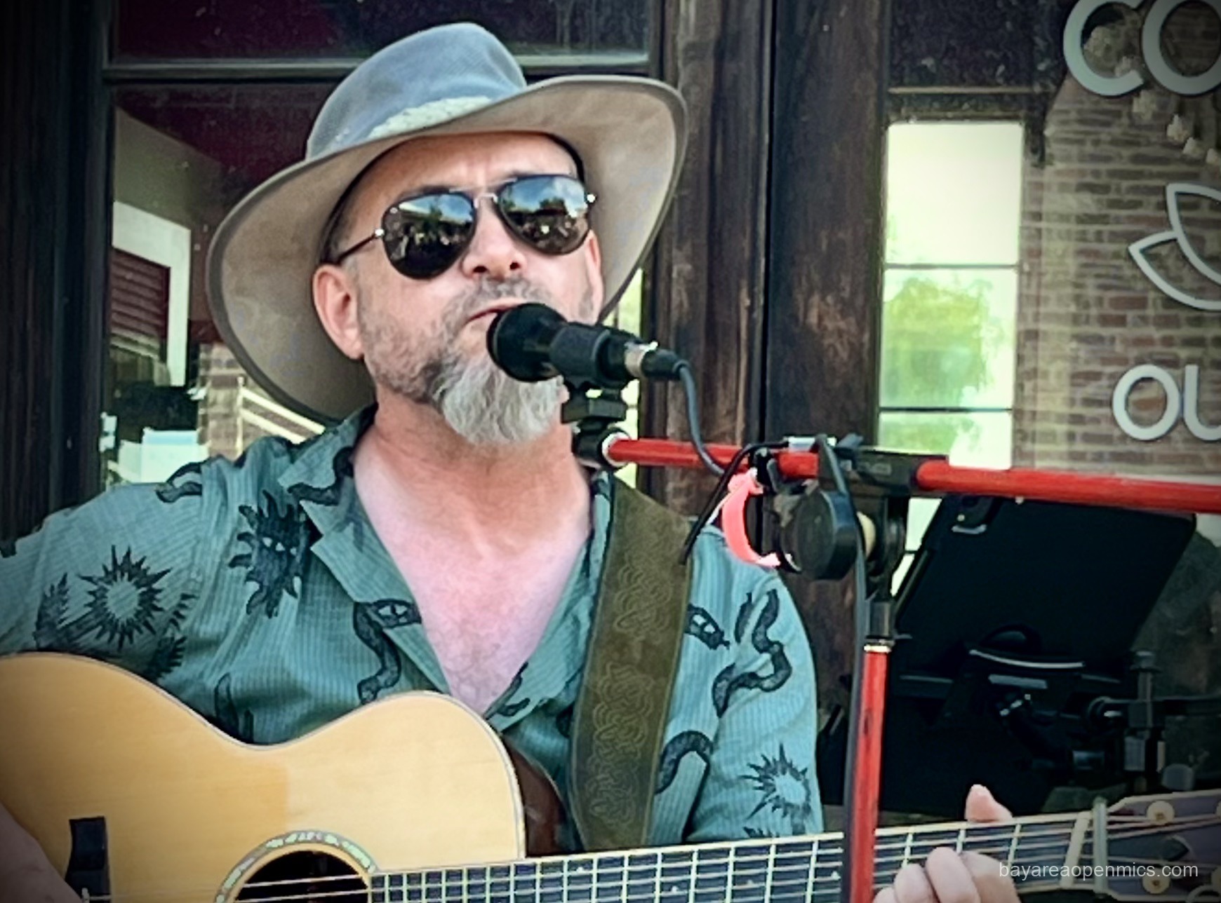 a man wearing sunglasses and broad-brimmed hat sings and plays guitar.