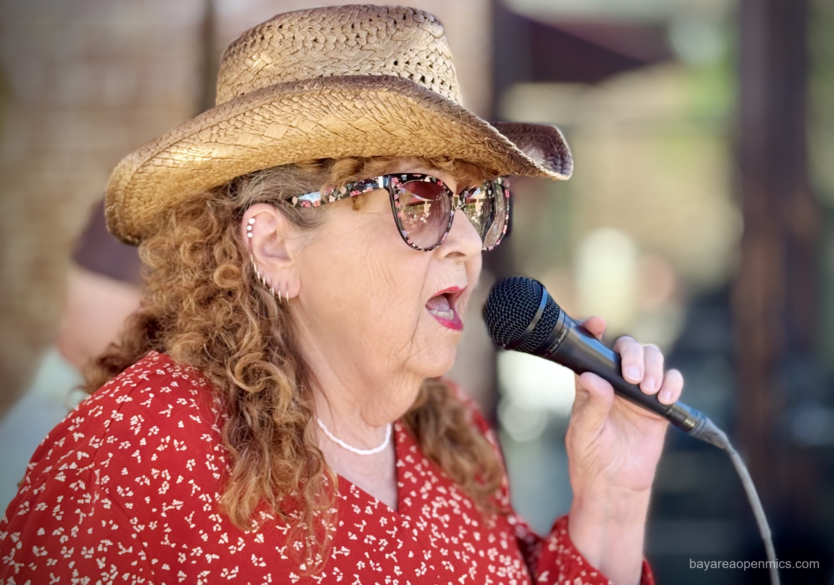 a woman in a red blouse, straw cowboy hat, and floral-print glasses hold a microphone while singing 