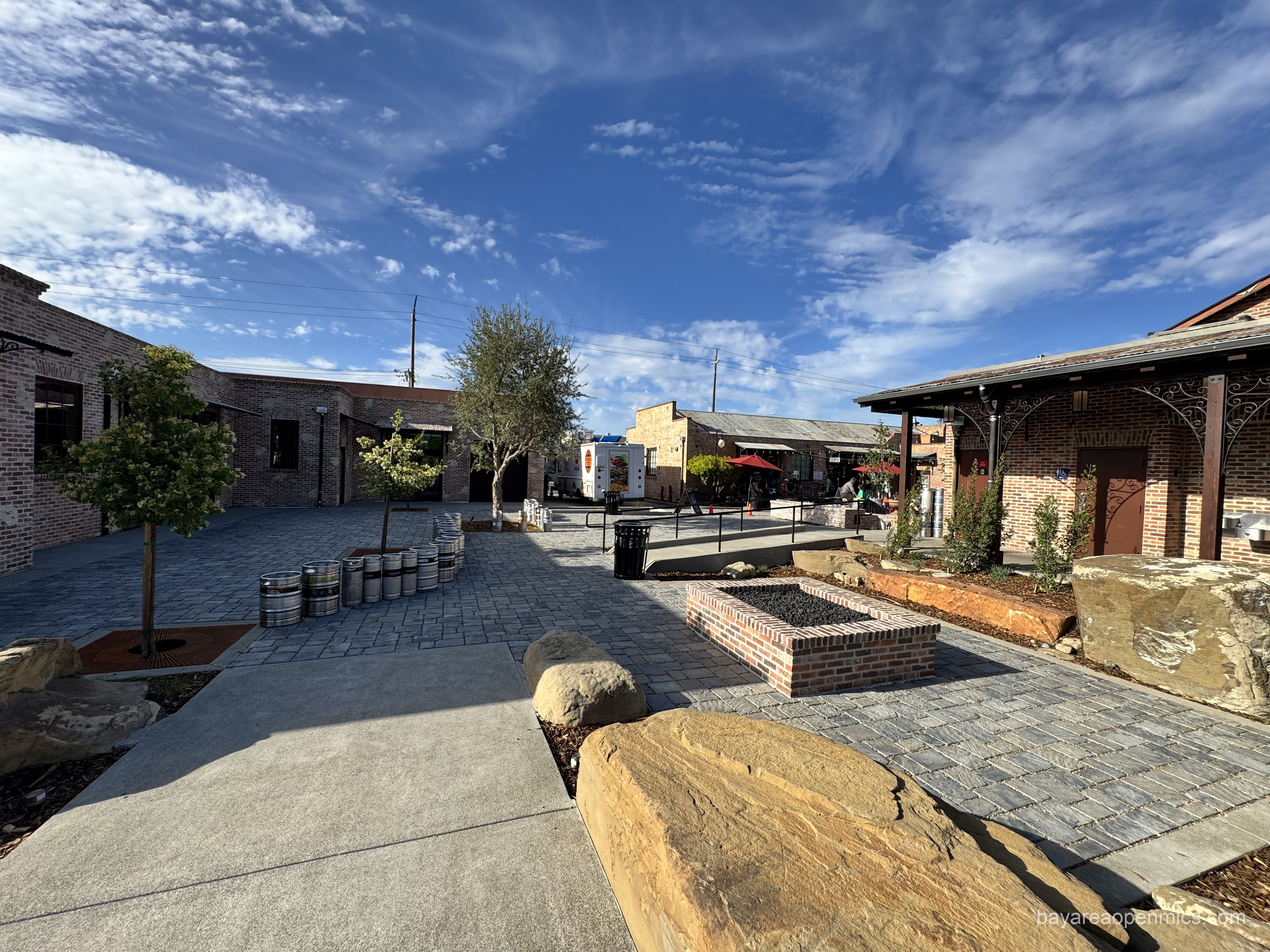 Large boulders, a rectangular brick firepit with black rocks, and a few new trees decorate a pavingstone courtyard between brick buildings A line of steeel beer kegs stretches along to the left.