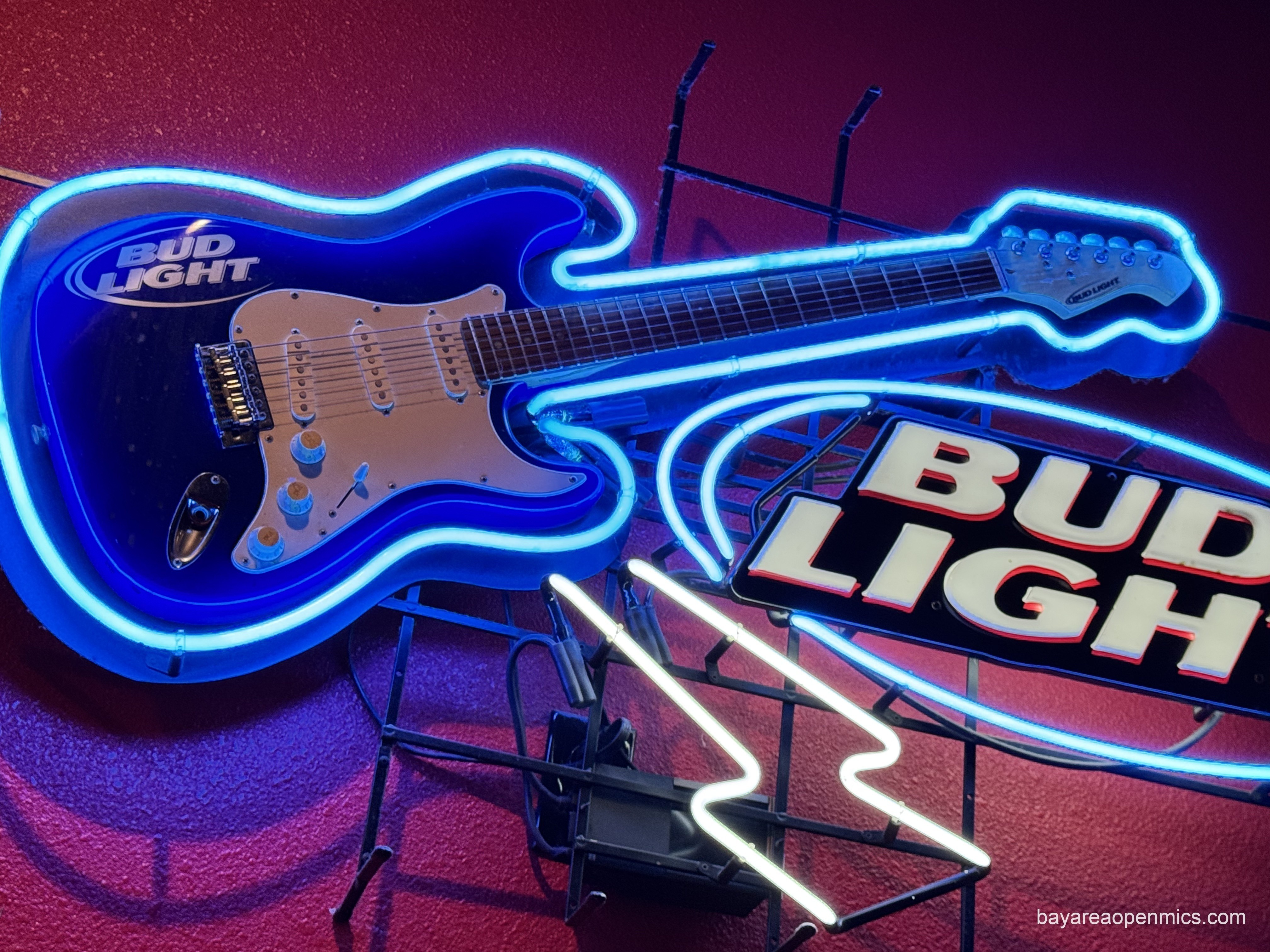 A blue stratocaster with a Bud Light logo on the body and headstock is mounted on a backing board and outlined in blue neon next to a larger, illuminated BUD LIGHT logo sign mounted on the wall