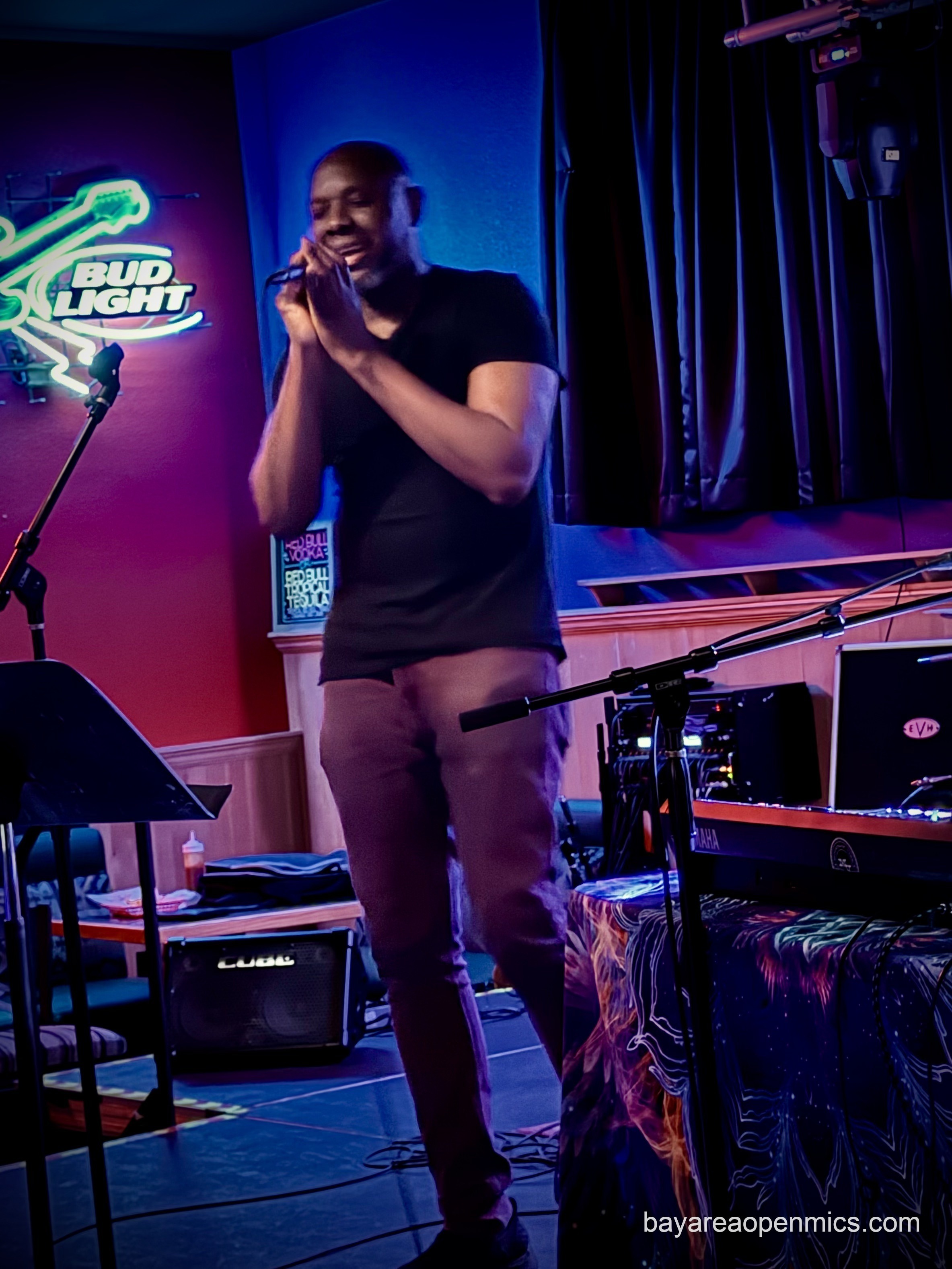 An African-American man with a bald head in a black t-shirt and brown pants holds a mic passionately while singing with his eyes closed