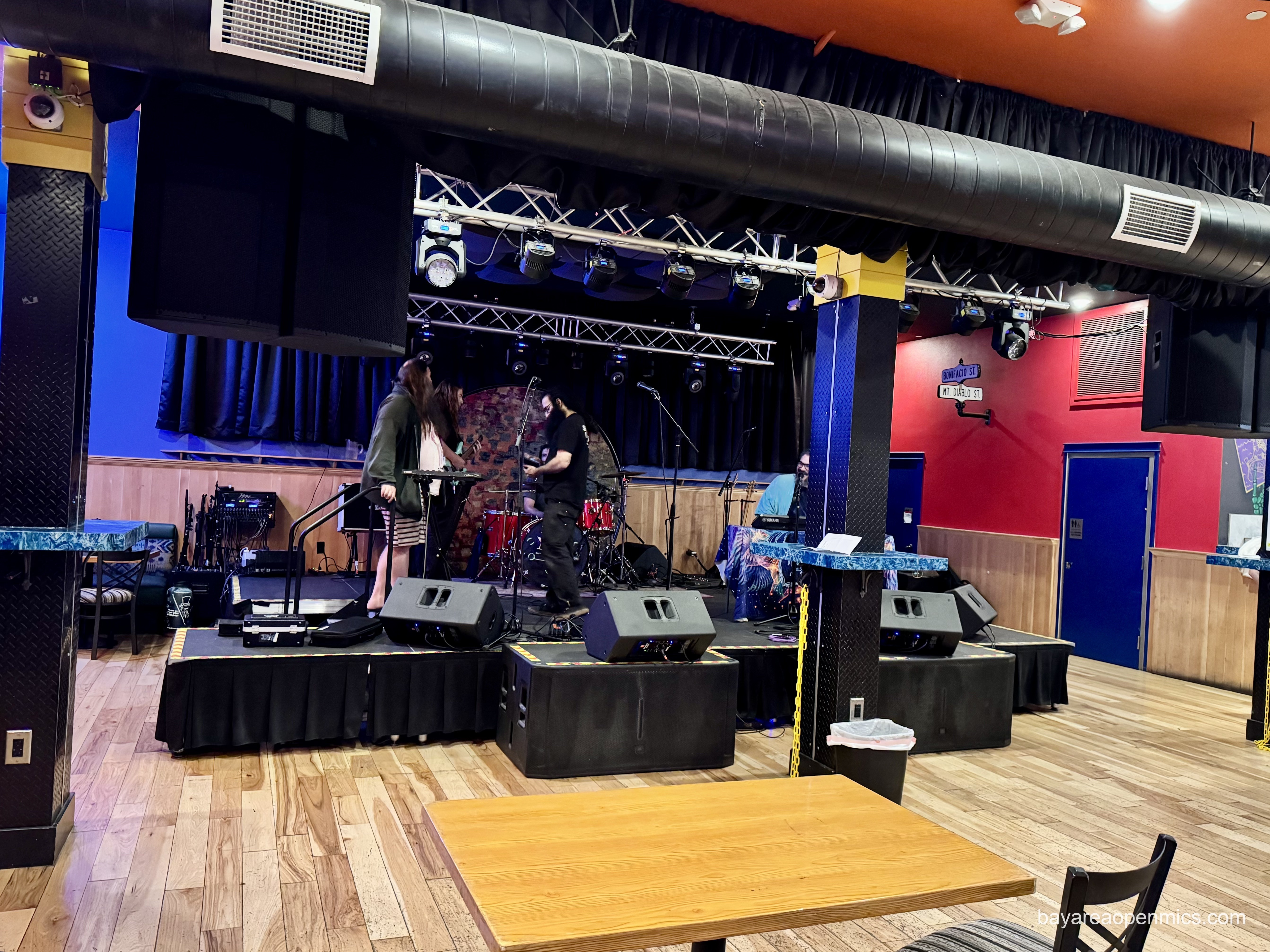 Brightly lit room with a large, professional stage on richly textured wooden floor with lights and speakers hung from the ceiling, men mill about the musical equipment