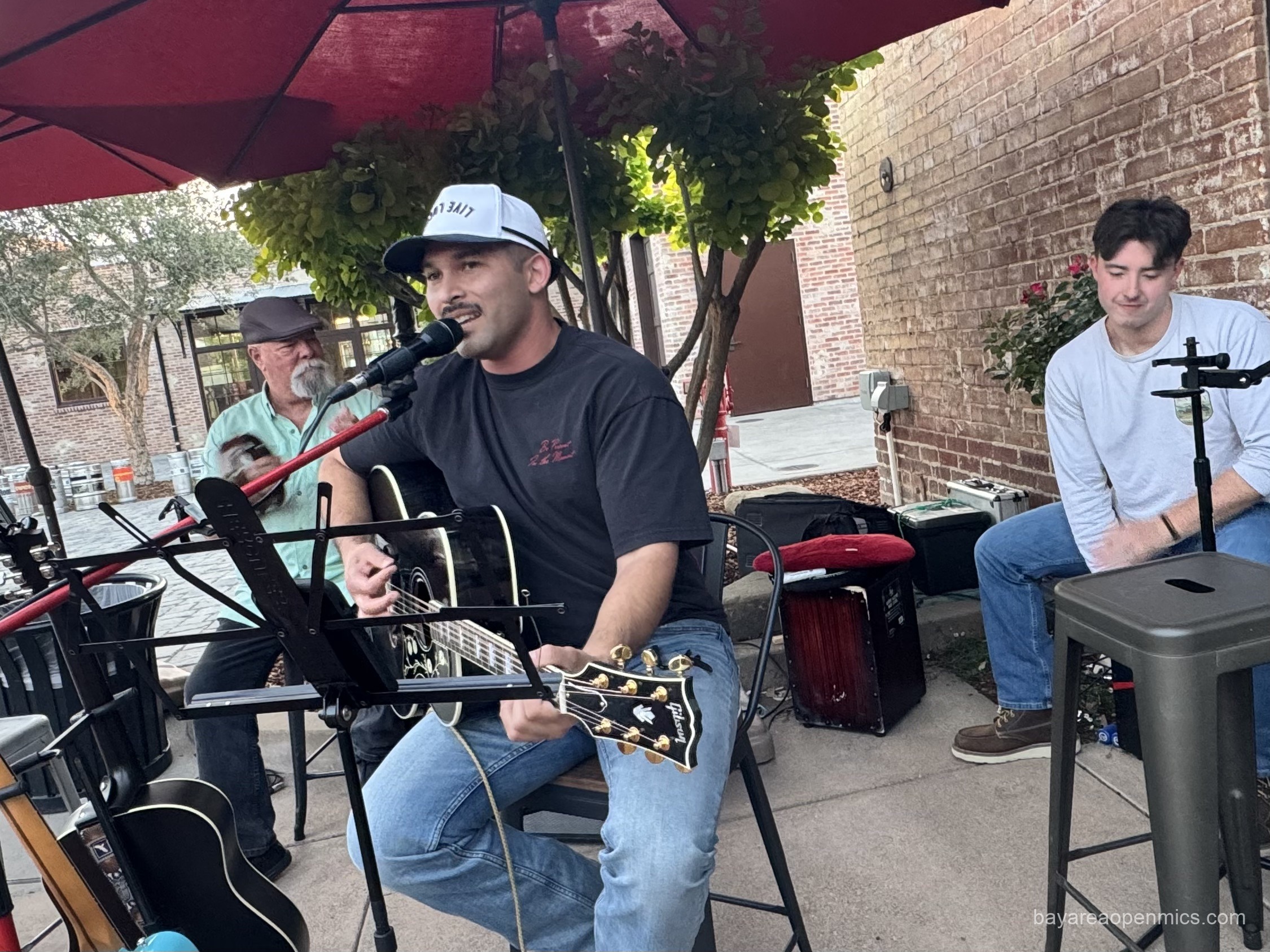 A smiling man with a thin mustache in a t-shirt and jeans plays a guitar and sings flanked by two percussionists: one shaking a tambourine to his right and the other plays a cajon to his left