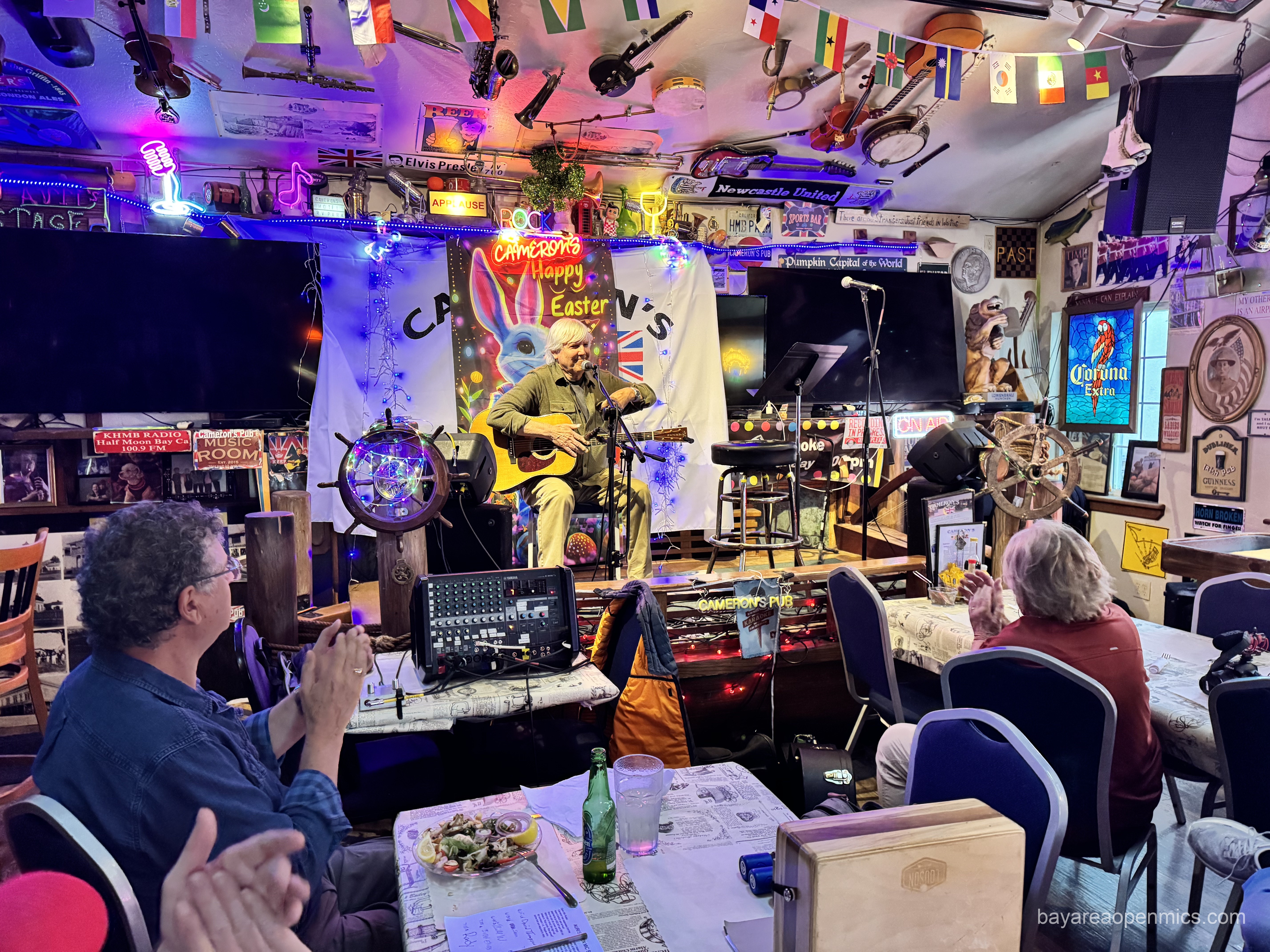 Seated people clap, facing the stage where a seated white-haired, clean-shaven man holding a guitar smiles. The room and its walls are cluttered with signs and bric-a-brac
