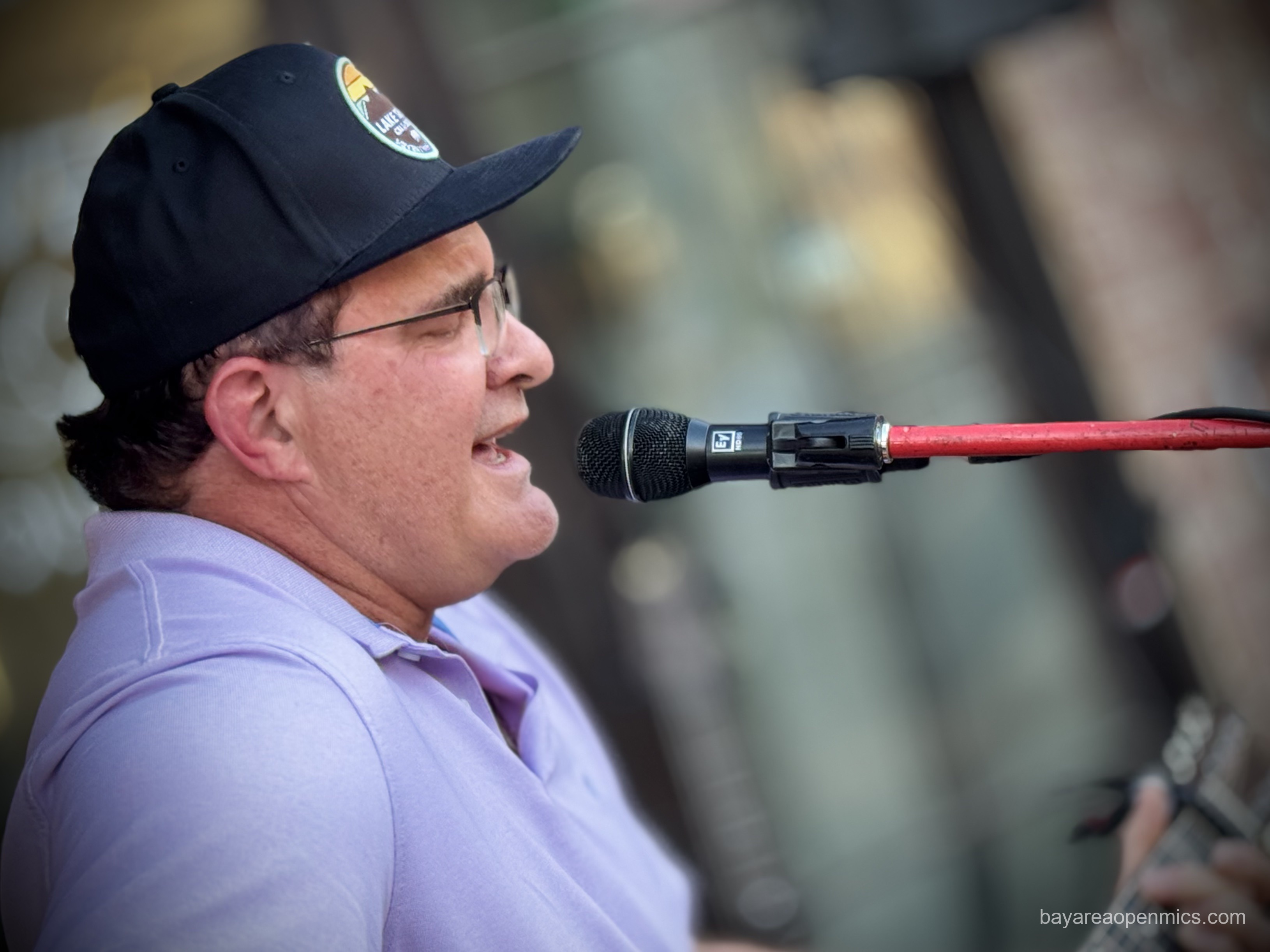 A man in a black baseball cap and glasses sings passionately into a microphone while strumming a guitar