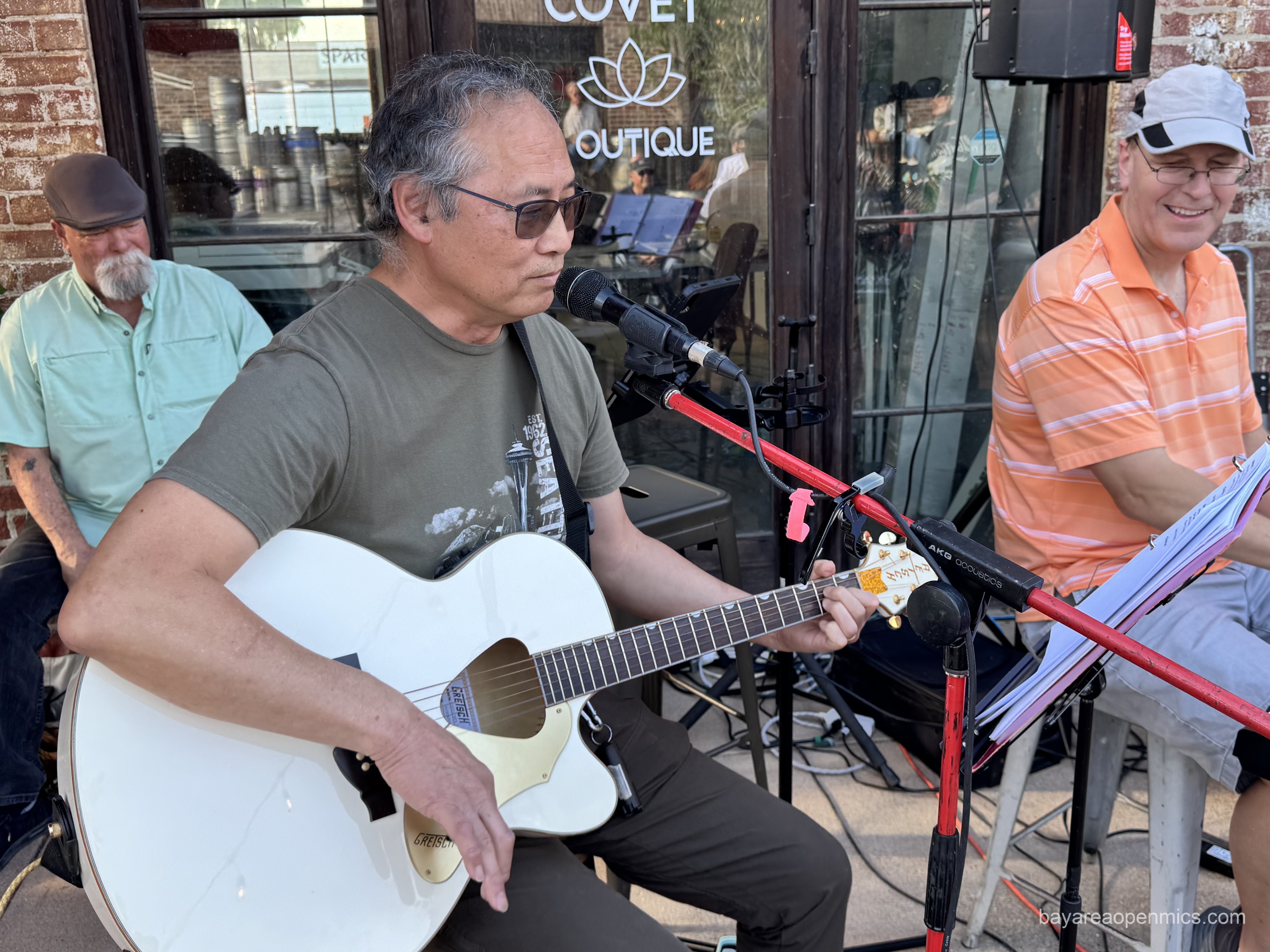 an asian man in glasses playing a white Gretch guitar with a triangular sound hole sits in front of a mic, a smiling man in a baseball cap and an orange shirt sits nearby on the keyboard, and third man in a flat cap, button up, with an extravagant goatee beard sits atop a cajon behind the first two men