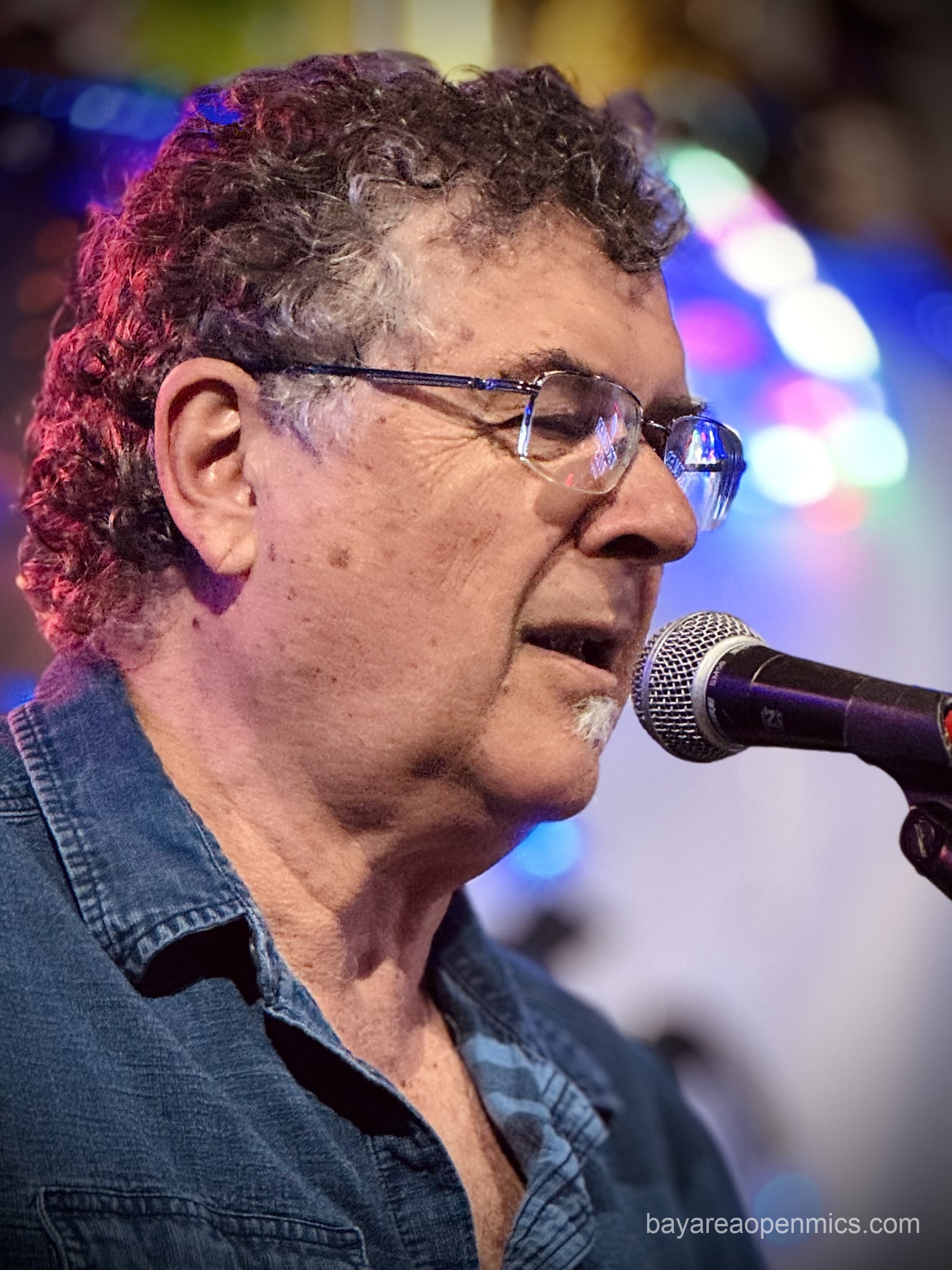 A curly-haired man in glasses and a jean button-up closes his eyes while singing in front of a microphone