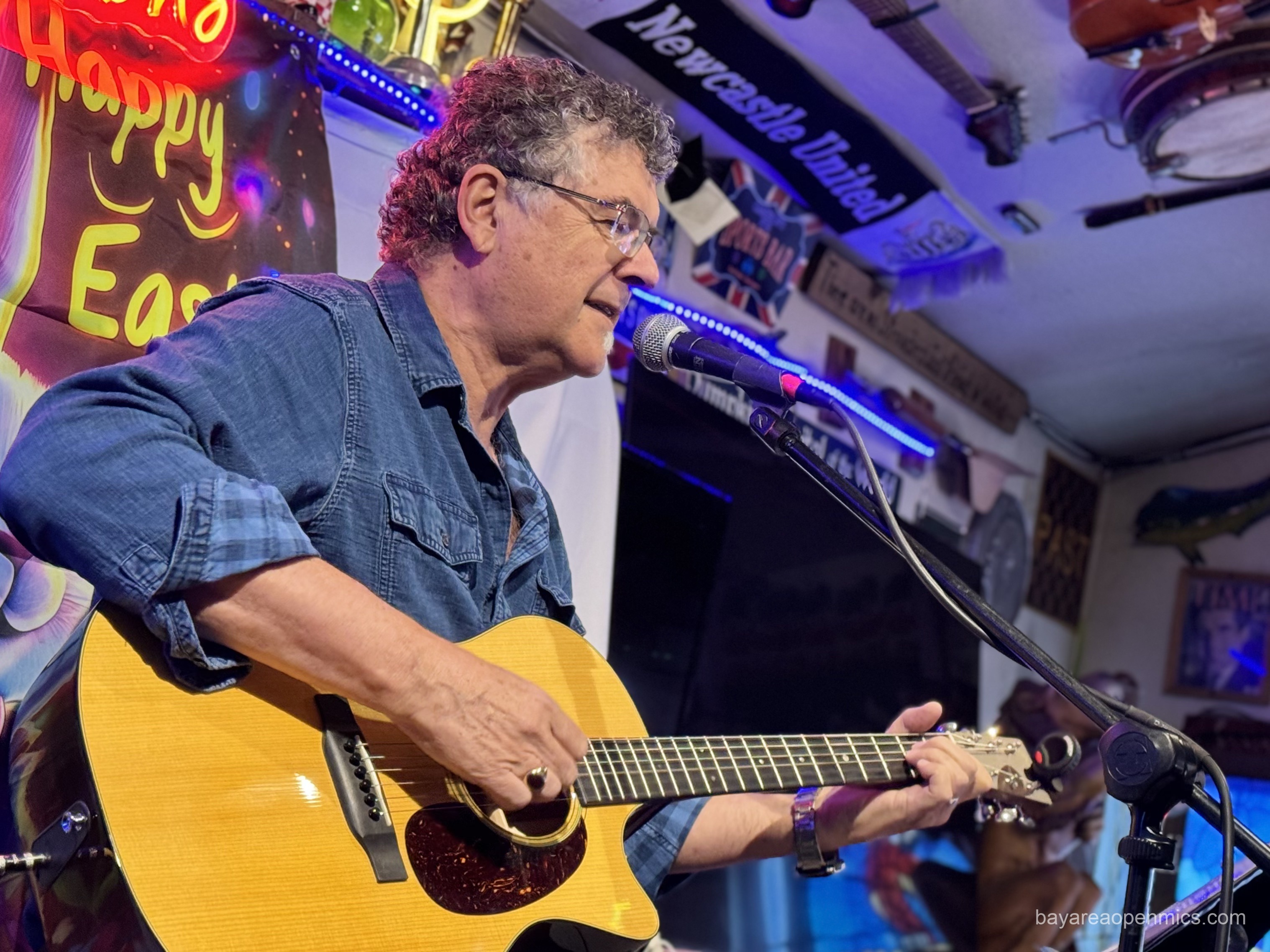A middle-aged man in glasses and a jean button-up plays a Taylor guitar while singing int a microphone