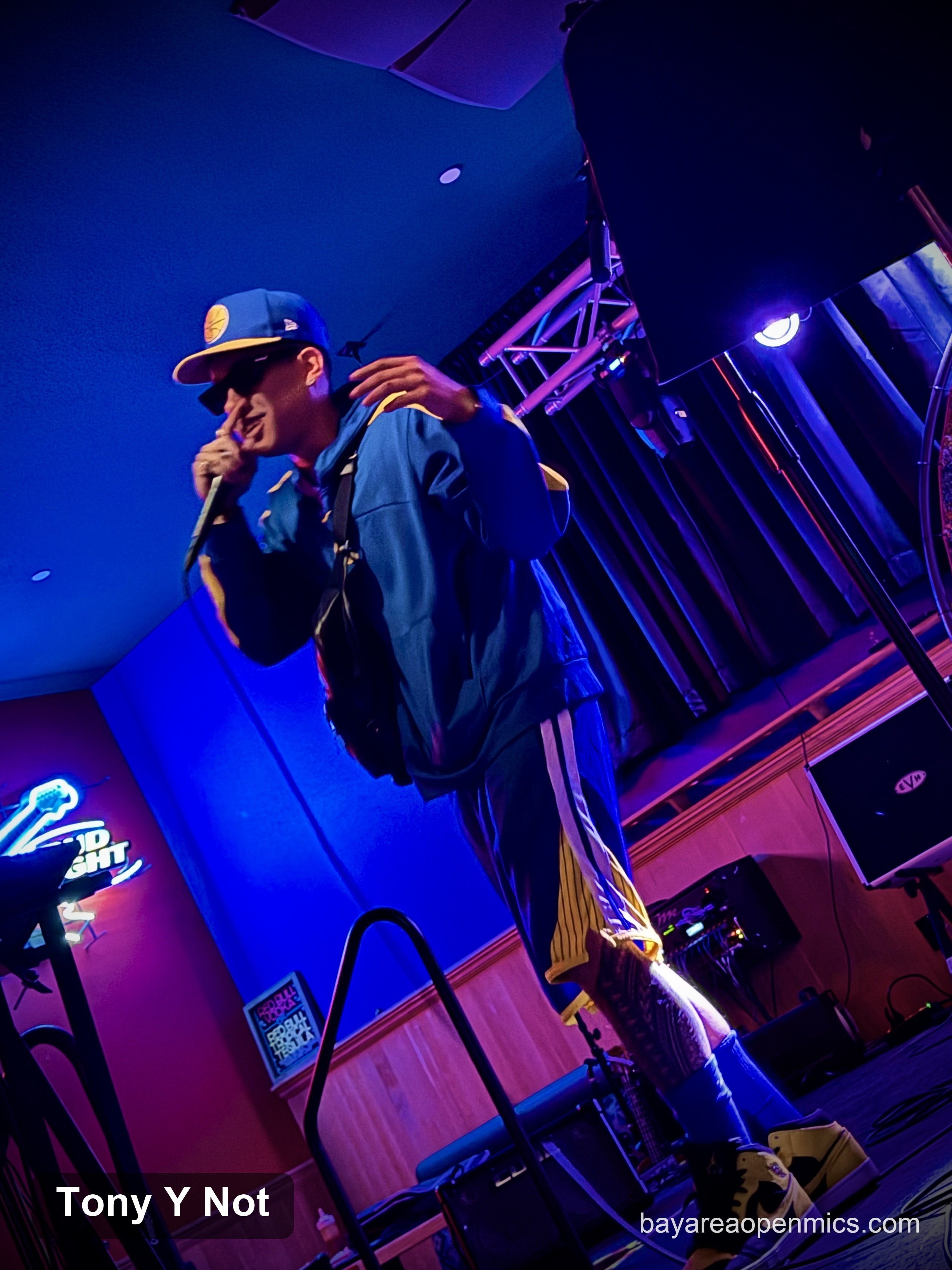 A rapper in sunglasses and a Golden State Warriors-branded baseball cap, jacket, basketball shorts, high socks, and shoes raps into a mic under the blue and pink lights