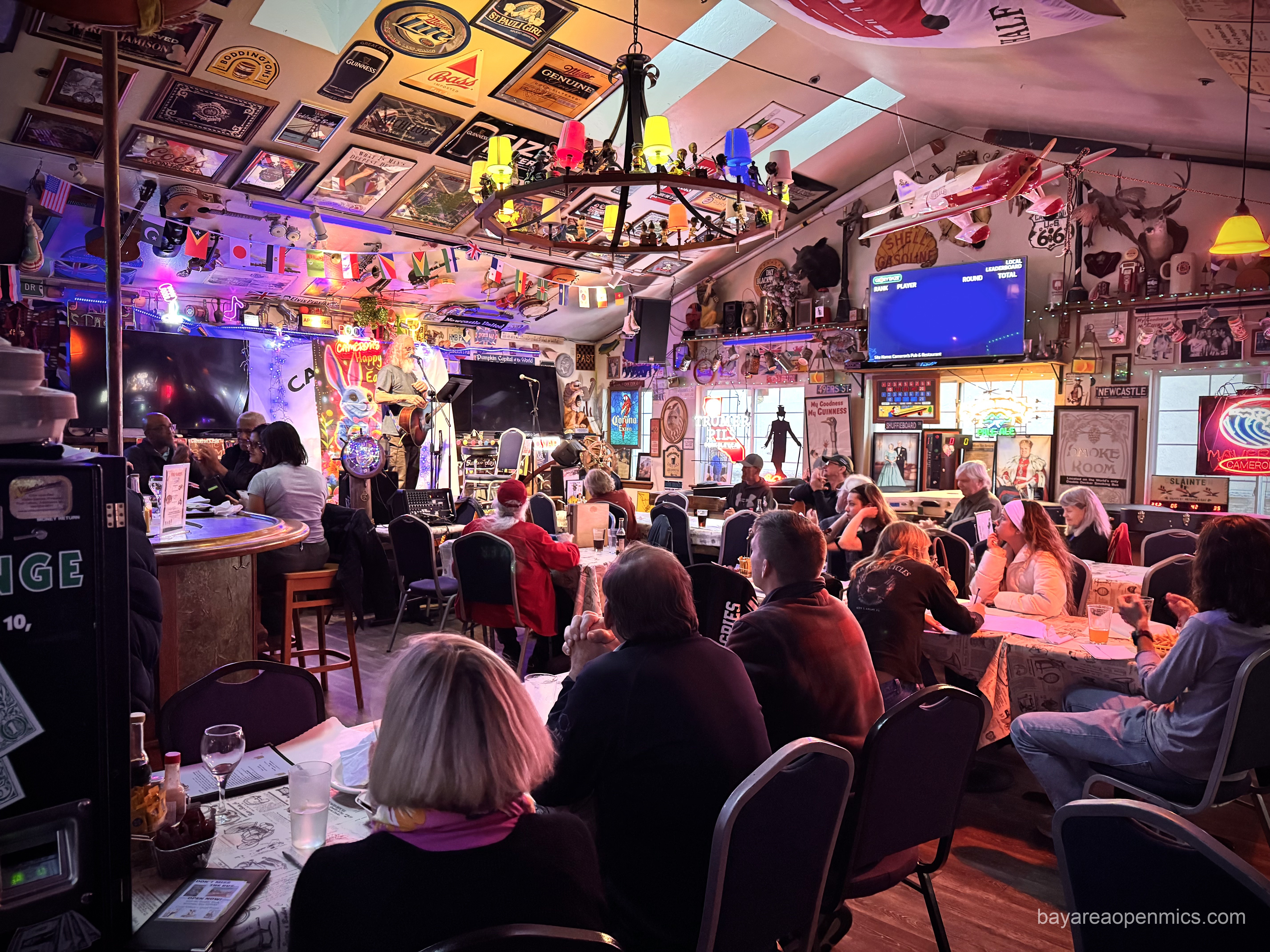 A crowd seated at tables before a stage with a standing singing guitarist