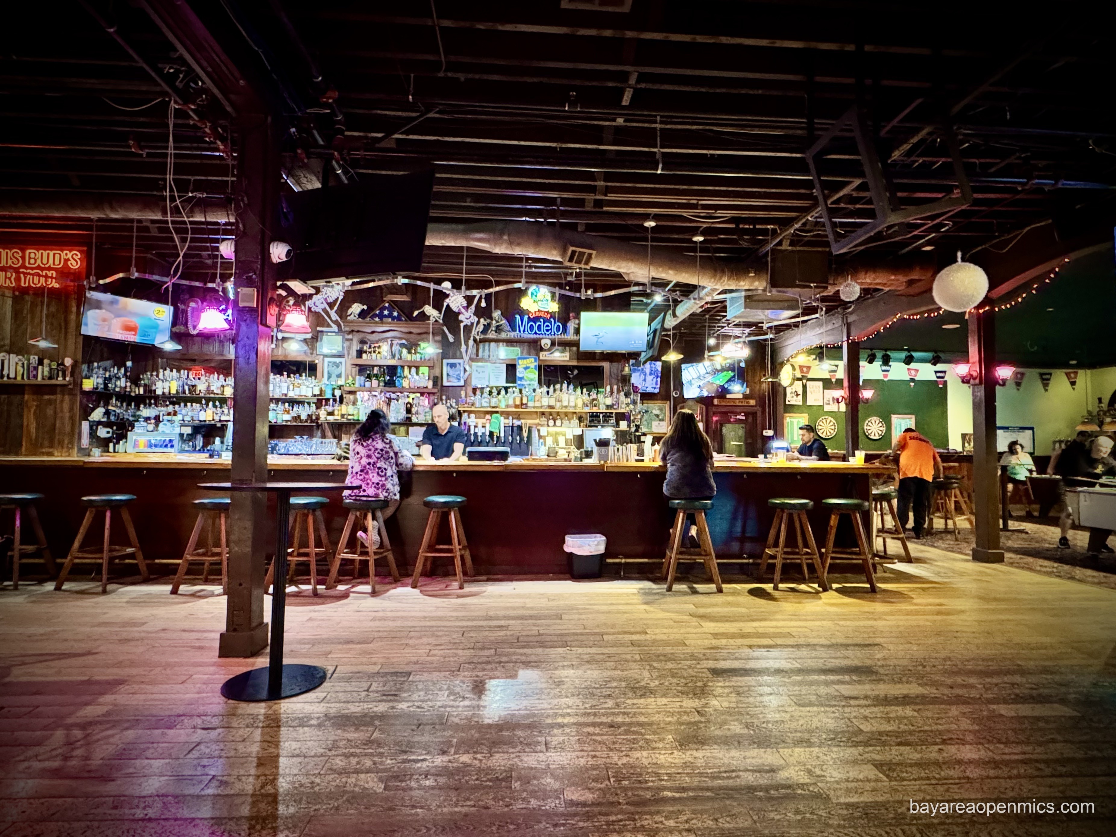 A cavernous room with wood floors shows a long bar with an impressive collection of bottles on the shelves behind it. In front there are ten stools with just a few patrons seated, spaced out from one another. A young, bald bartender is behind the bar, seemingly chatting with a patron