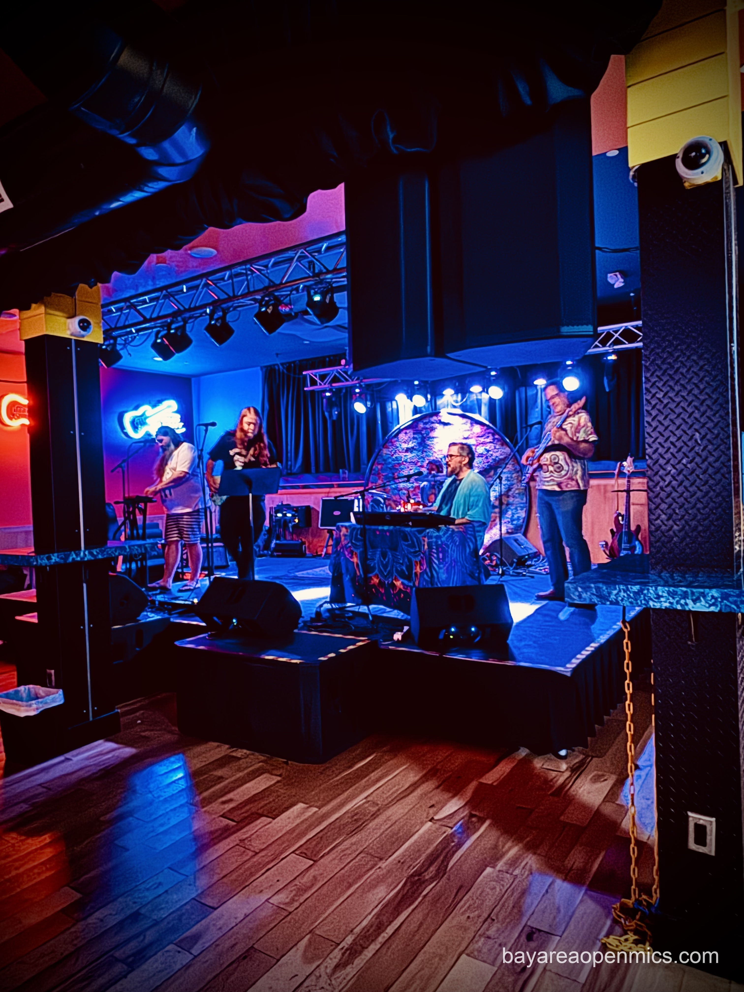 Massive speakers both on a richly textured wooden floor and hung from the ceiling frame five musicians under colorful blue and red onstage lights: from the left, a long-haired man in a t-shirt and striped shorts plays a sampler pad; another long-haired man plays a headless electric guitar while looking down at a music stand; Joey Nunez, a bespectacled short-haired man with a goatee, sings while playing keyboard; behind Joey a drummer is partially seen; and on the right of the photo, a tall bass man in jeans and a t-shirt with short hair and glasses plays a six-string electric bass guitar
